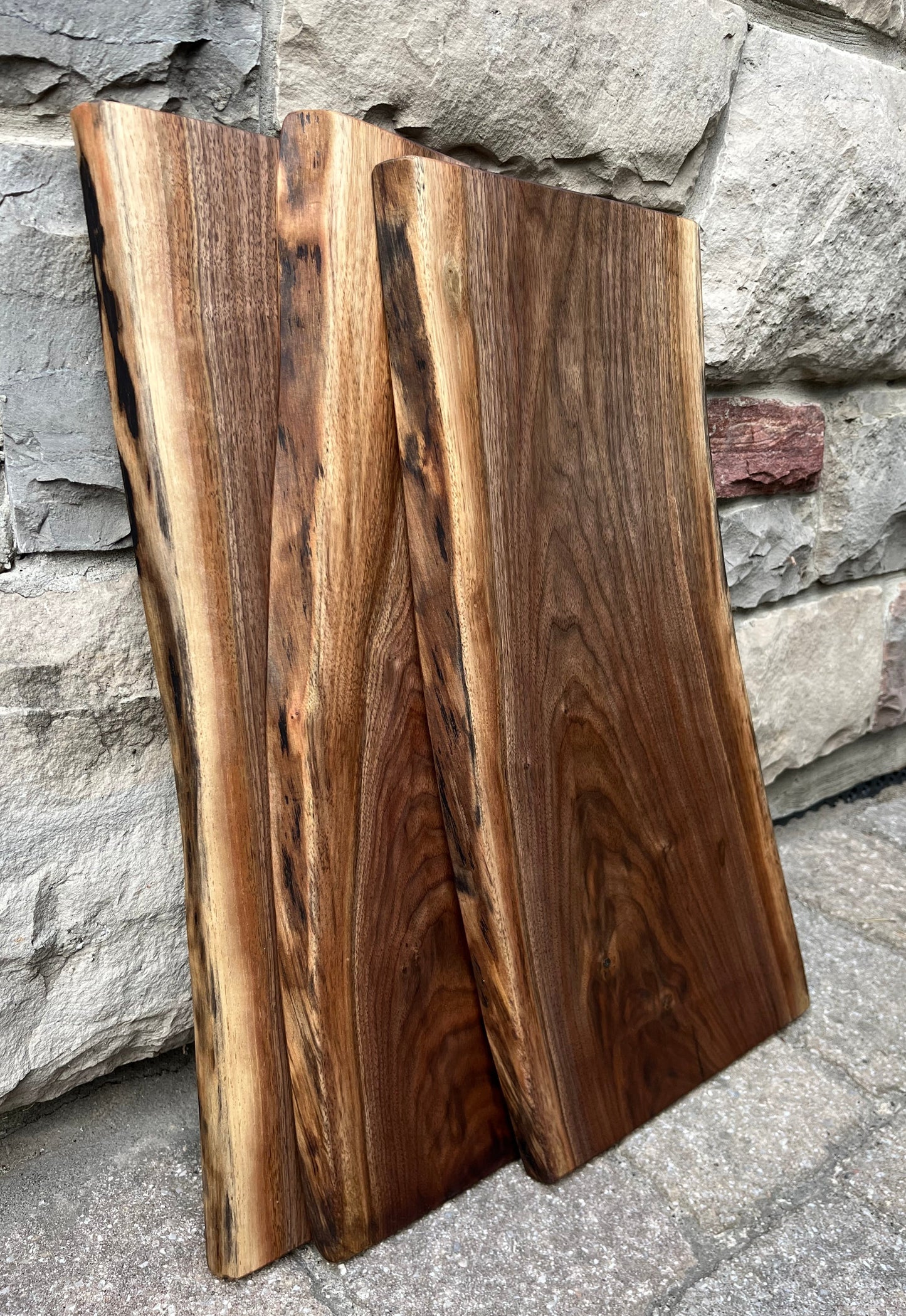 Three 22-inch long and 8-12-inch wide live-edge black walnut charcuterie boards leaning against a stone wall