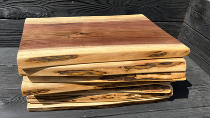 side angle of seven 16-inch long and 8-11-inch wide live-edge black walnut charcuterie boards stacked up with black wood panel background