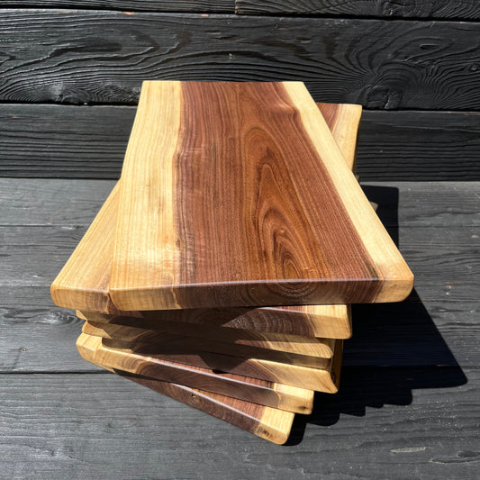 Seven 16-inch long and 8-11-inch wide live-edge black walnut charcuterie boards stacked up with black wood panel background