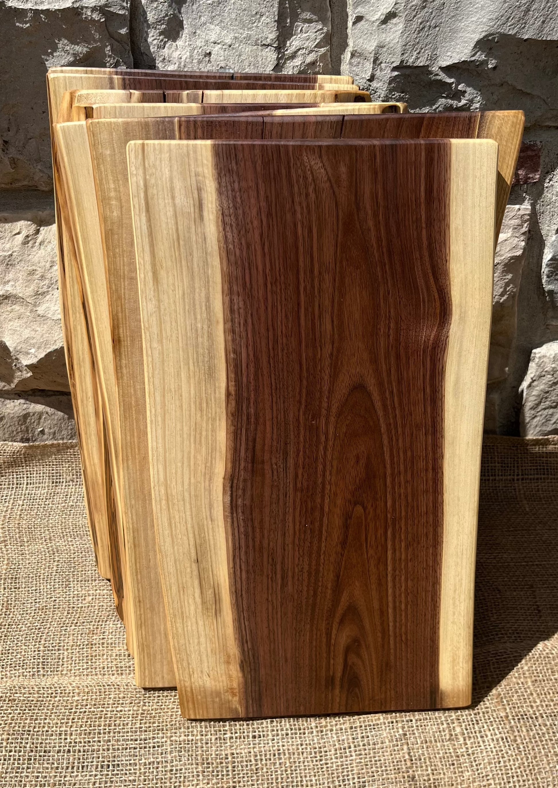 front face view of seven 16-inch long and 8-11-inch wide live-edge black walnut charcuterie boards stacked up against a stone wall background