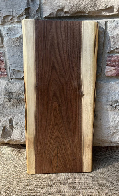 Single 22-inch long and 8-12-inch wide live-edge black walnut charcuterie board leaning against a stone wall on a burlap base