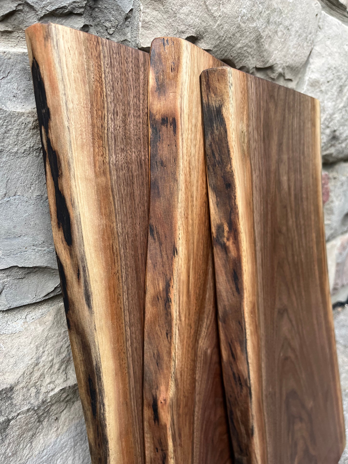 Side live-edge view of three live-edge black-walnut charcuterie boards leaning against a stone wall