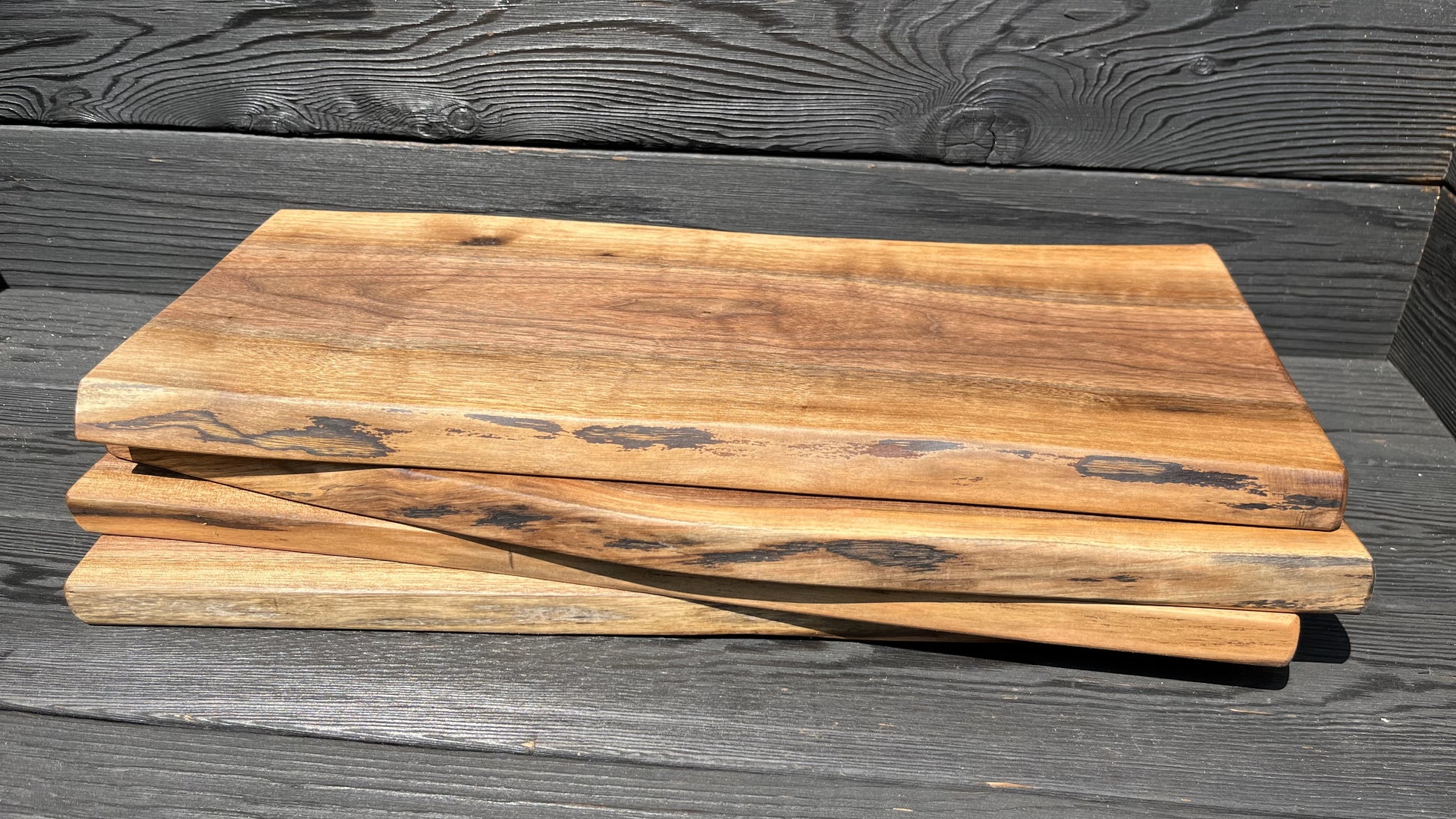 Side view of four 22-inch long and 8-12-inch wide live-edge black walnut charcuterie boards stacked up with black wood panel background