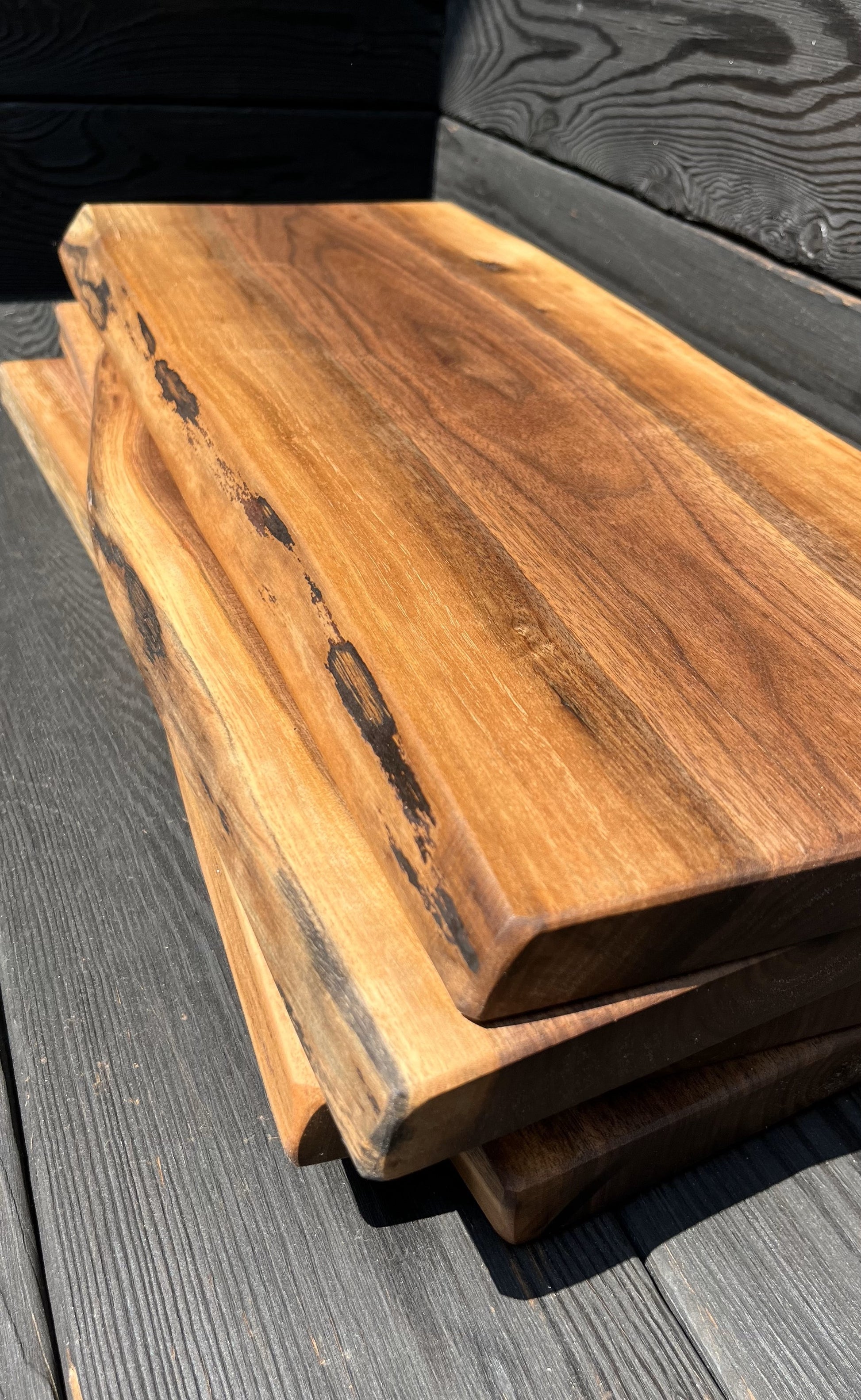 Four 22-inch long and 8-12-inch wide live-edge black walnut charcuterie boards stacked up with black wood panel background